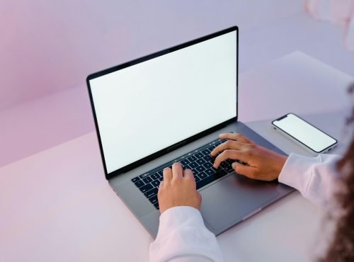 A person working on a laptop with a blank screen and smartphone on a desk.